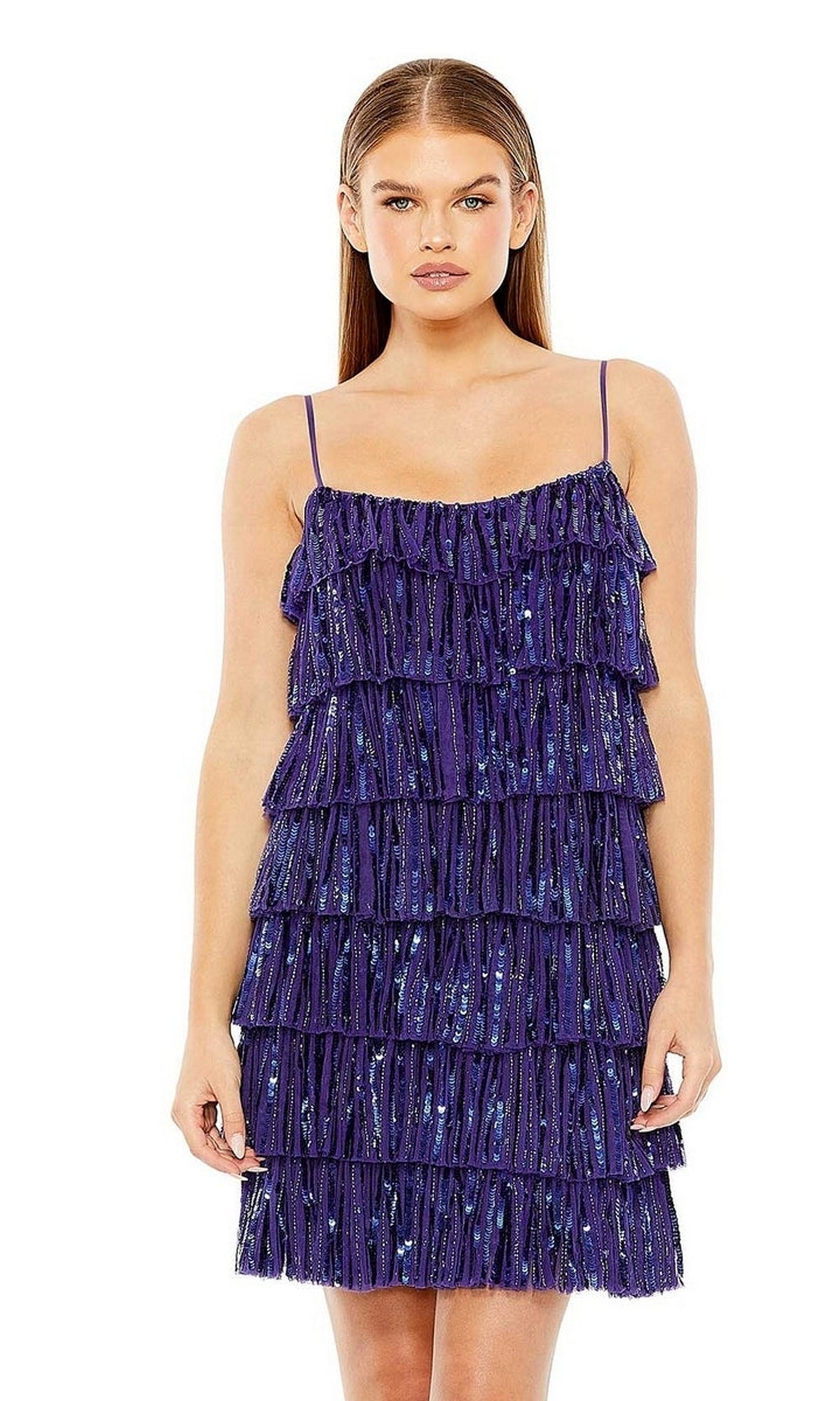 A woman stands facing forward, wearing the Mac Duggal 23001—a short ruffled semi-formal dress with thin straps in deep blue, featuring tiered fringe and sparkling sequin accents.